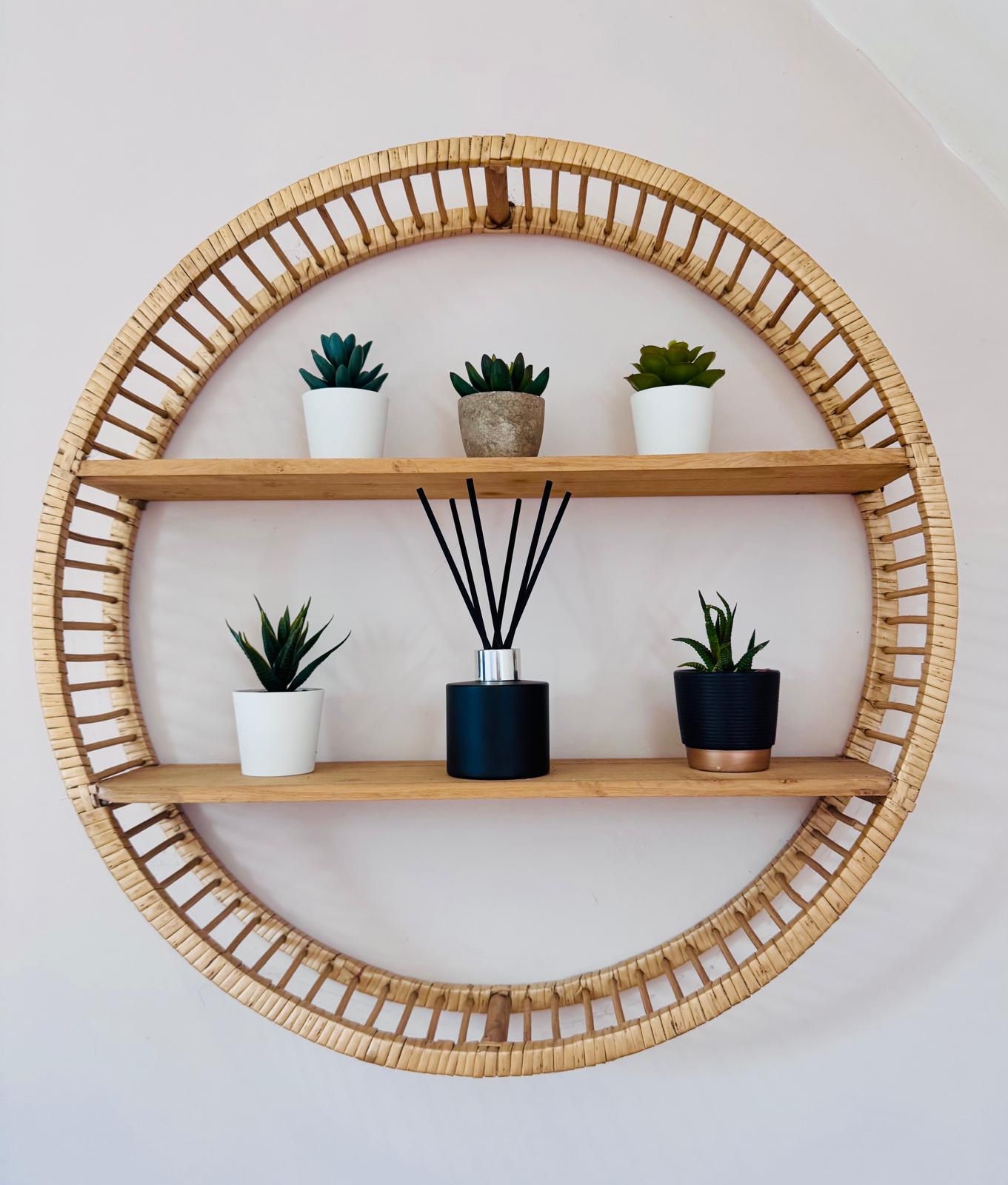 Round wooden shelf with wicker frame holding plants and a diffuser on a white wall.