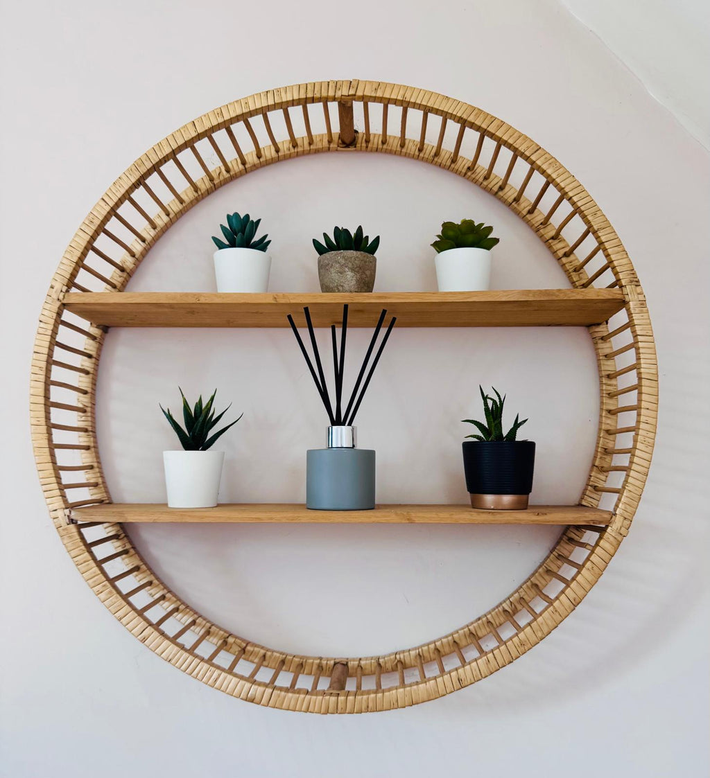 Round wooden shelf with wicker frame holding plants and a diffuser on a white wall.