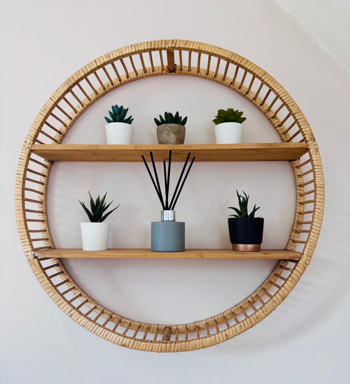 Round wooden shelf with wicker frame holding plants and a diffuser on a white wall.