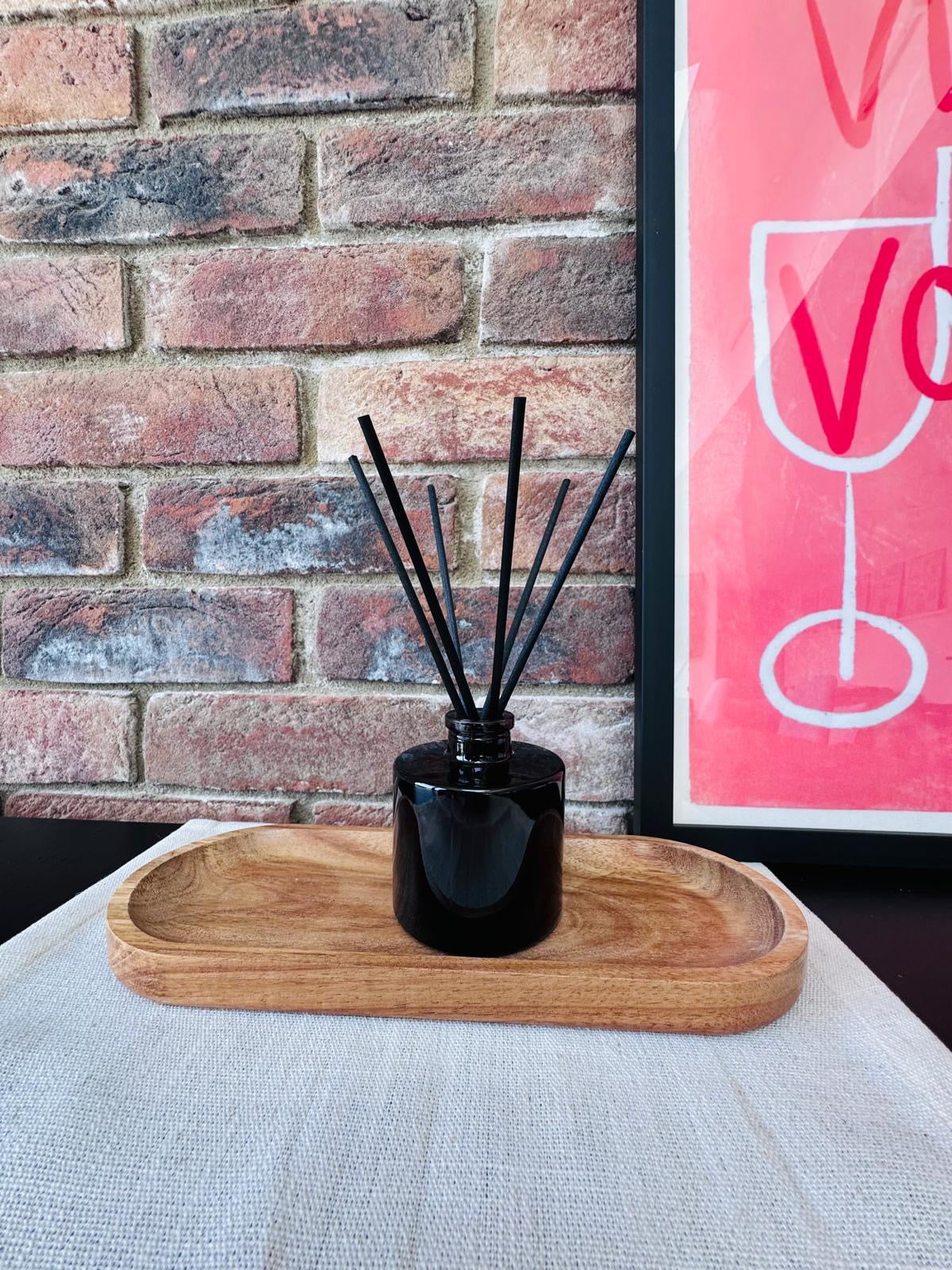 Black diffuser with reeds on a wooden tray against a brick wall with a pink sign.
