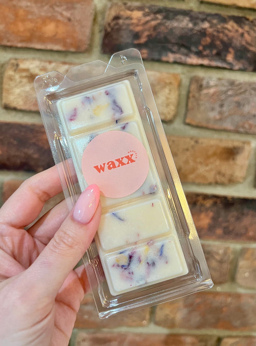 Packaged wax melts with 'waxx' branding held against a brick wall.